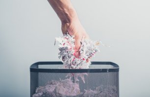 A hand is putting a bunch of shredded paper in a waste paper basket