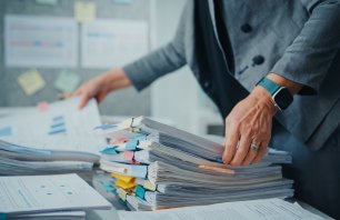 Close-up of overworked mature businesswoman manager sorts through a huge stack of files and documents at cluttered desk in office. Struggling to keep up with daily paperwork and deadlines.